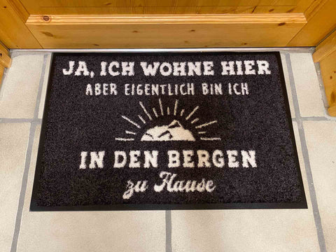 Schwarze Fussmatte mit weissem Text "In den Bergen zu Hause" und Berg-silhouette Widderstein