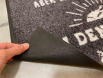 Detail schwarze Fußmatte mit weißem Text, Hand zeigt Gummi-Rückseite